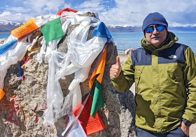 Traveler photo: A guest stands before prayer flags by Namtso Lake, capturing a photo with the colorful flags and sacred water behind. (August 2026)
