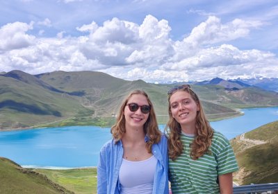 Traveler photo: Two guests smile side by side by Yamdrok Lake, with blue water and distant mountains framing their natural group shot. (August 2026)