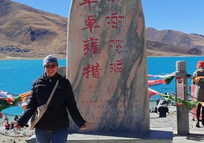 Traveler photo: By elevation marker stones beside Yamdrok Lake in Tibet, our guest with bright smile against sapphire waters, instantly frozen in time. (February 2026)