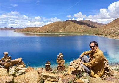 Traveler photo: Mani stones pile up along the sacred lake as the guest smiles into the frame, merging faith with the landscape. (July 2026)