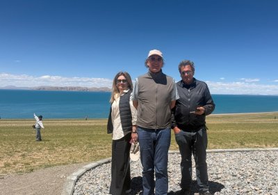 Traveler photo: Three guests line up at Namtso Lake, smiling toward the camera with turquoise water and mountains behind. (July 2026)